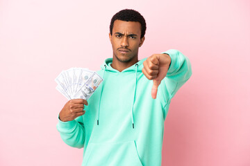 Young man taking a lot of money over isolated pink background showing thumb down with negative...
