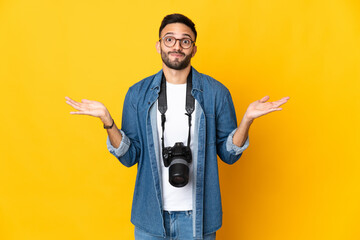 Young photographer girl isolated on yellow background having doubts while raising hands