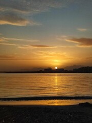 Beautiful sunset over the calm sea, with silhouettes of buildings and distant mountains. Golden sunlight reflects on the water, creating a tranquil scene