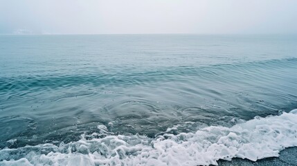 Fototapeta premium Gentle waves kiss the foggy shoreline, merging sea and sky into a seamless expanse of tranquil, misty blues.