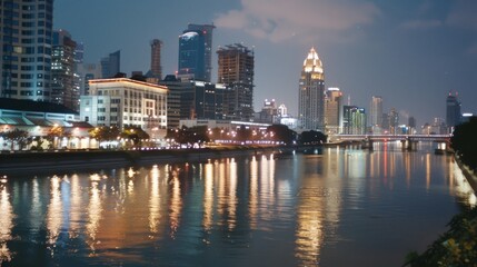 A tranquil riverside scene captures the reflection of city lights shimmering across the water, as modern skyscrapers stand tall under the night sky.