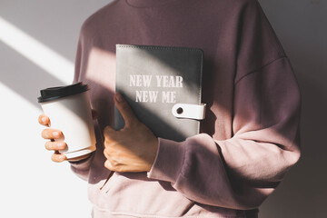 female hand holding a grey diary with the text new year new me and paper cup with hot drink on the...
