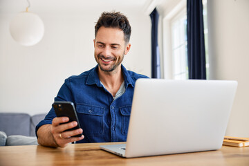 Cheerful man using his phone while working from home on his laptop