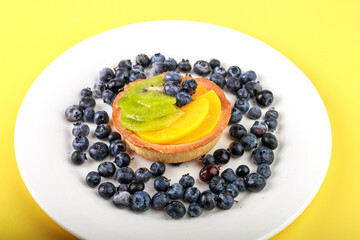 shortbread basket cake with cream, blueberries, peaches and kiwi on a plate on a yellow background