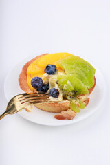 fork breaking shortbread basket with cream, blueberries, peaches and kiwi on a plate on a white background