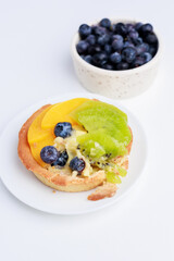 fork breaking shortbread basket with cream, blueberries, peaches and kiwi on a plate on a white background