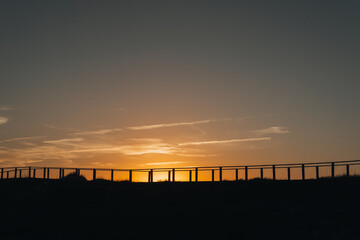  A minimalistic sunset scene featuring a silhouetted boardwalk against a golden sky. The calm, open landscape evokes a sense of peace and quiet at day's end