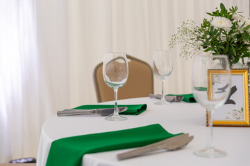 Close up of wine glasses, silver tableware and green napkins on round tables covered with white tablecloth.