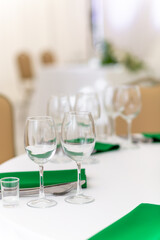 Close up of wine glasses, silver tableware and green napkins on round tables covered with white tablecloth.