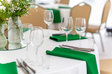 Close up of wine glasses, silver tableware and green napkins on round tables covered with white tablecloth.