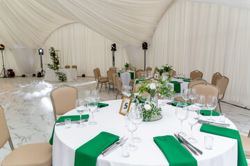 Wine glasses, silver tableware and green napkins on round tables covered with white tablecloth.