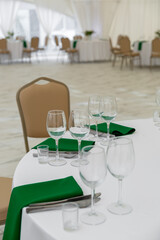 Close up of wine glasses, silver tableware and green napkins on round tables covered with white tablecloth.