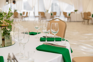 Close up of wine glasses, silver tableware and green napkins on round tables covered with white tablecloth.