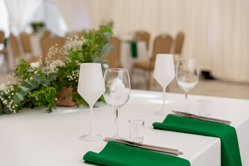Close up of wine glasses, silver tableware and green napkins on round tables covered with white tablecloth.