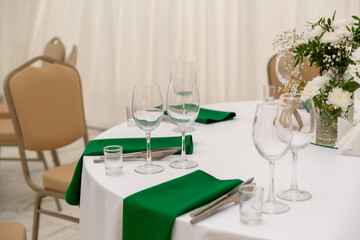 Close up of wine glasses, silver tableware and green napkins on round tables covered with white tablecloth.
