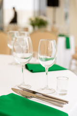 Close up of wine glasses, silver tableware and green napkins on round tables covered with white tablecloth.