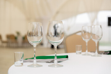 Close up of wine glasses, silver tableware and green napkins on round tables covered with white tablecloth.