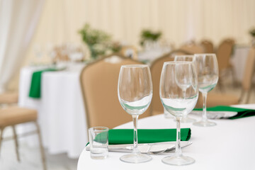 Close up of wine glasses, silver tableware and green napkins on round tables covered with white tablecloth.
