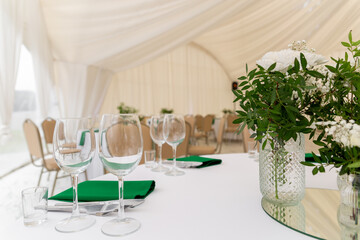 Close up of wine glasses, silver tableware and green napkins on round tables covered with white tablecloth.
