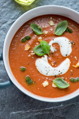 Roasted tomatoes and red bell pepper soup topped with cream, basil pesto and green basil, vertical shot, middle close-up, elevated view