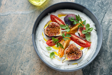 Bowl of ricotta dipping sauce with fresh figs and tomatoes, horizontal shot on a beige and grey granite background, high angle view