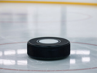 A detailed close-up of a black hockey puck resting on the ice surface of a rink, highlighting the texture and design of the puck against the smooth ice. Winter sports.