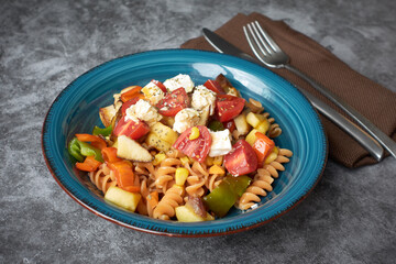 Blue ceramic plate with fresh pasta salad with cherry tomatoes, feta cheese pieces and vegetables like pepper, corn and apple. Fork and knife on brown napkin on concrete or stone background