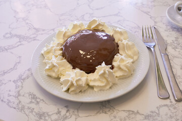 A decadent dessert covered in rich chocolate, surrounded by swirls of whipped cream on a white plate. A fork and knife are set beside it, ready to enjoy this indulgent treat