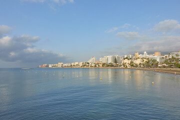 Naklejka premium Calm coastal cityscape of Benalmadena Costa, village of Malaga, Andalusia, Spain. Blue water and beachside buildings under a partly cloudy sky, showing a serene Mediterranean shoreline