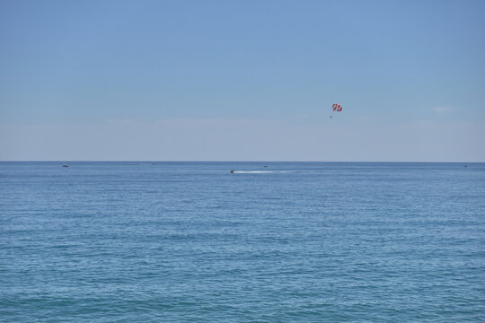 A serene seascape featuring calm blue waters under a clear sky, with a distant parasailing activity visible as a small red dot, creating a peaceful Mediterranean summer atmosphere