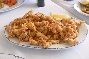 A generous portion of golden fried squid or calamari served on a white decorative plate with lemon wedge, ready to share on a white tablecloth in a restaurant setting