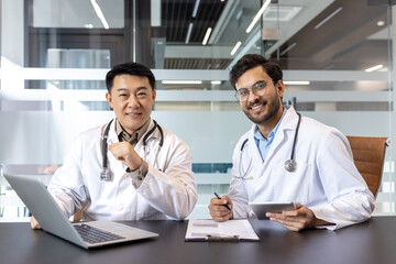 Two medical professionals in white coats collaborate in modern office. One smiles using laptop, other holds tablet. Emphasizing teamwork, technology, healthcare in professional setting.