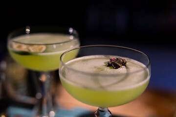 Glass of gin based Basil Smash cocktail on bar. Two cocktails of gin. Blurry background. Basil smash alcoholic cocktail drink with dry gin, syrup, lemon, basil and ice, dark bar counter background.