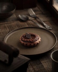 A single chocolate tart with a crispy crust and a swirl of chocolate filling sits on a brown plate. A small piece of chocolate sits on top.