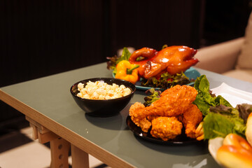 The cozy Christmas party setup features variety of beautifully wrapped gift boxes on table, alongside festive food dishes fried chicken, grilled chicken, salad, popcorn, and bread in living room.