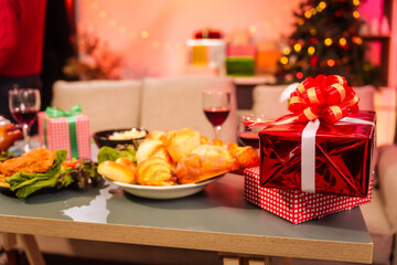 The cozy Christmas party setup features variety of beautifully wrapped gift boxes on table, alongside festive food dishes fried chicken, grilled chicken, salad, popcorn, and bread in living room.