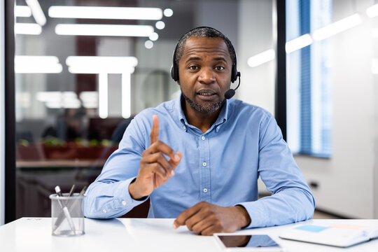 Mature african american businessman wearing headset in modern office during video conference. Engaging in discussion with expressive gestures, focused on communication and teamwork.
