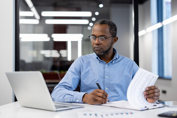 Mature African American businessman in modern office working on laptop, reviewing documents....