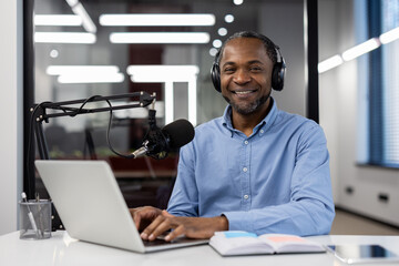 Mature African American businessman records business course video using laptop and microphone. Wearing headphones, he smiles confidently in office. Educational, professional atmosphere depicted.