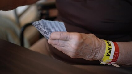 Closeup of an elderly persons hands holding playing cards, with a yellow Fall Risk wristband visible on their wrist. - Powered by Adobe