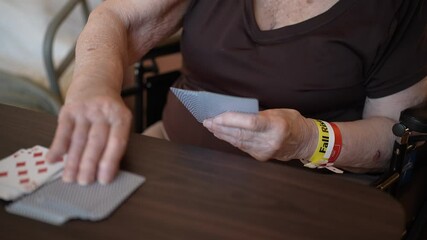 Closeup of an elderly persons hands holding playing cards, with a yellow Fall Risk wristband visible on their wrist. - Powered by Adobe