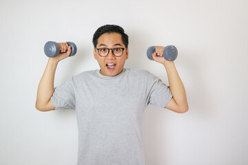 An Asian man smiles confidently while holding two blue dumbbells in his upright position, wearing a gray t-shirt. He stands against a white background in a fitness studio.