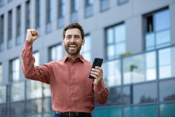 Mature man celebrating success outside, holding smartphone with lifted arm, expressing joy and...