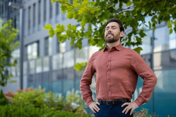 Mature man standing outside in urban area, breathing deeply with eyes closed. Wearing rust shirt,...