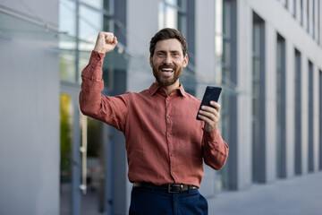 Confident mature man in business attire celebrates success outdoors with fist raised, holding smartphone. Captures a triumphant moment against modern urban backdrop.