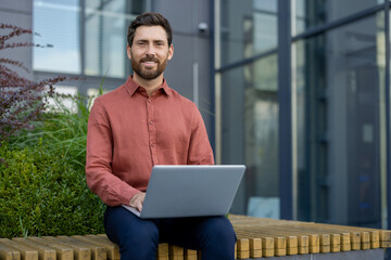 Mature man sitting on outdoor bench in front of modern office building, working on laptop with a smile. Casual yet professional, he reflects confidence and technology use.