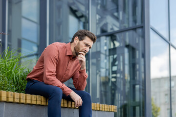 Thoughtful mature man in casual clothes sits outdoors, contemplating life decisions. Urban setting...