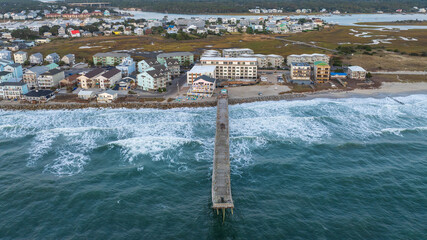 Aerial view of the pier at Carolina Beach, North Carolina, stretching into the Atlantic Ocean, with surrounding coastal homes and marshland in the background.
