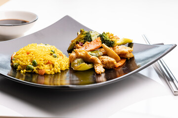 Szechuan chicken served with a scoop of fried rice on a black plate, accompanied by chopsticks and a small bowl of soy sauce.