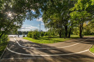 A serene park pathway curved along the riverside with walking inhabitants enjoying nature on a sunny afternoon.